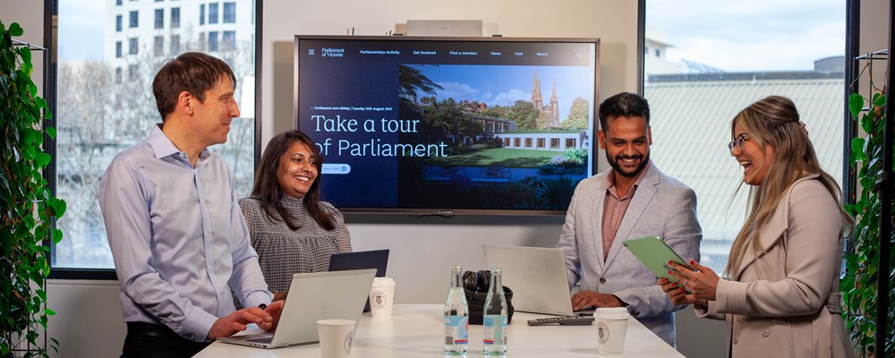 Four Parliament employees meet with a screen in the background with an image of Parliament 