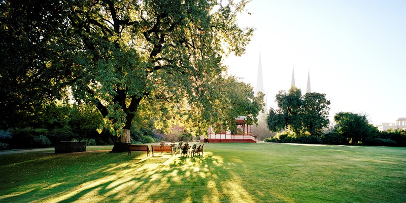A misty early morning on a green lawn in the garden. Chairs and tables are grouped under a large tree. Sunlight is streaming through the branches and casting shadows on the grass. A pavilion can be seen in the background, and in the distance there is three church spires.